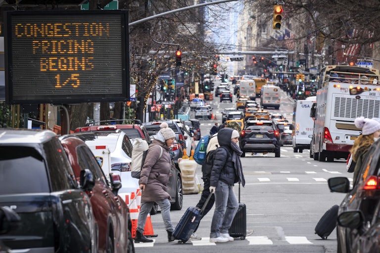 A group of people walk in New York City while a traffic sign reads "Congestion Pricing Begins 1/5."