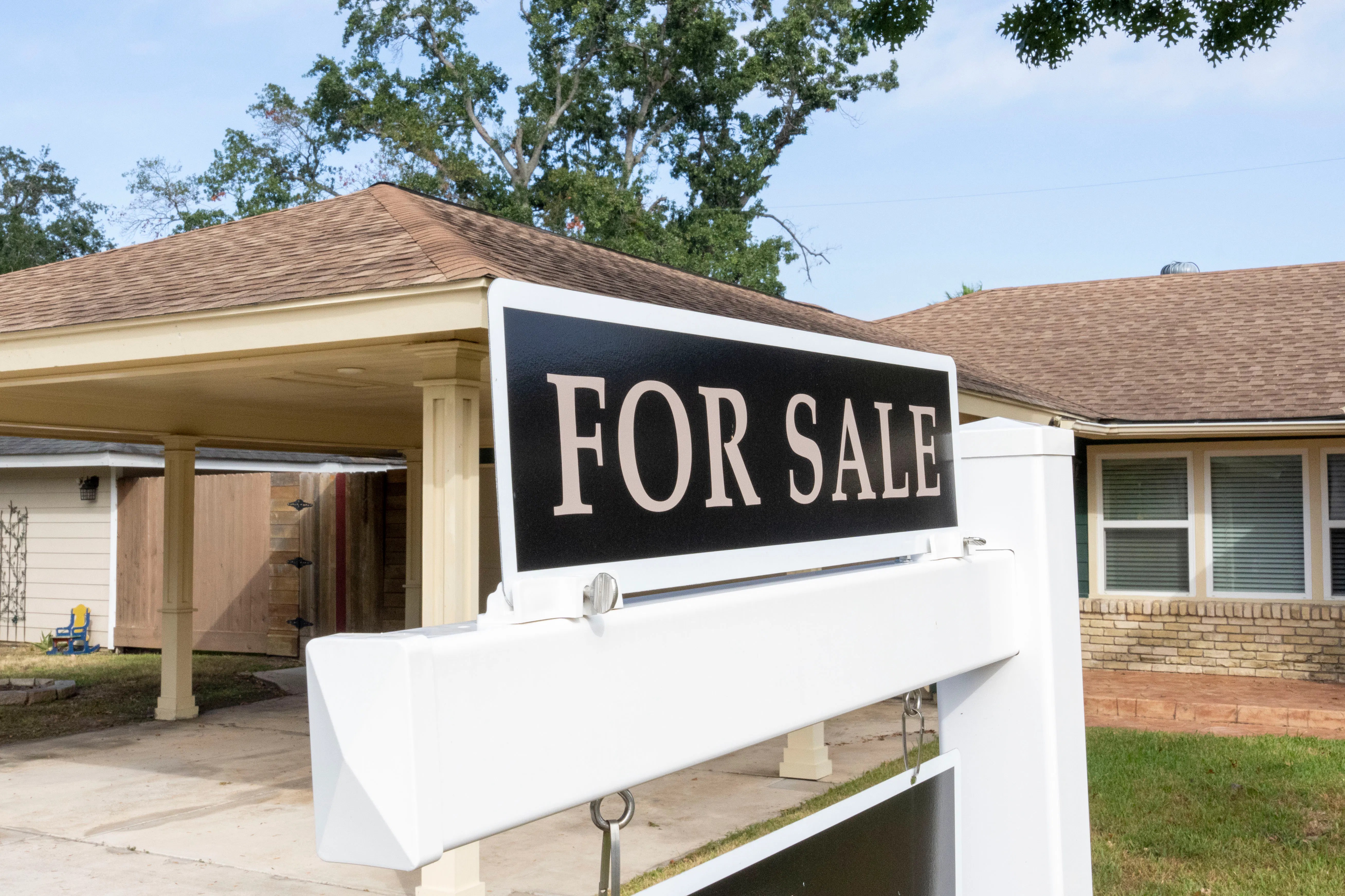 A "for sale" sign sits outside of a house in the daytime, with a driveway, car port, and trees visible behind it. 