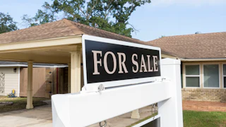 A "for sale" sign sits outside of a house in the daytime, with a driveway, car port, and trees visible behind it.