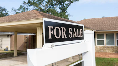 A "for sale" sign sits outside of a house in the daytime, with a driveway, car port, and trees visible behind it.