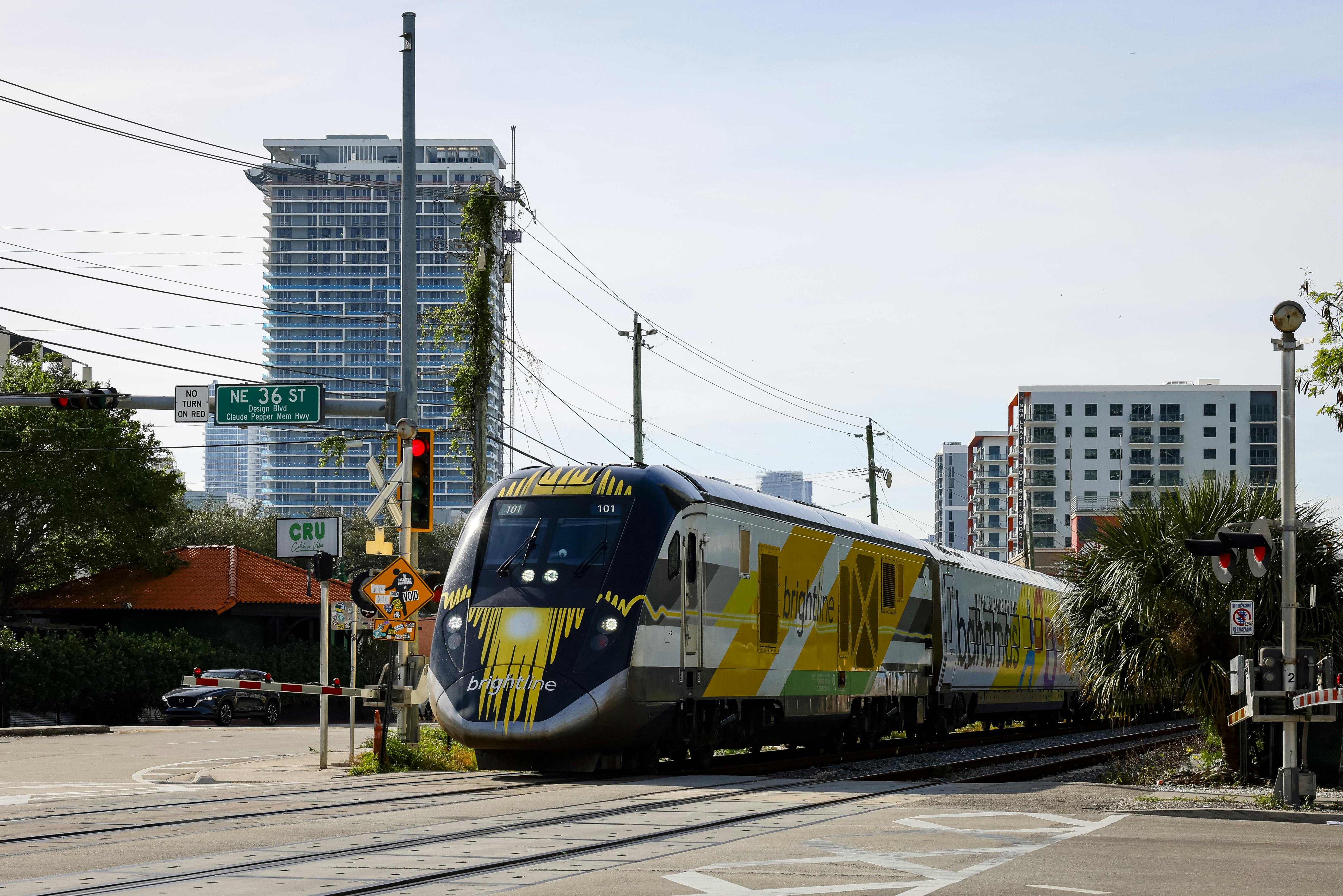 A Brightline train stands at an open, urban railway crossing.