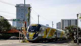 A Brightline train stands at an open, urban railway crossing.