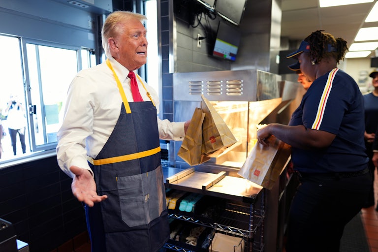 Donald Trump wearing a McDonald’s apron and holding two brown McDonald’s paper bags. Two McDonald’s employees stand nearby.