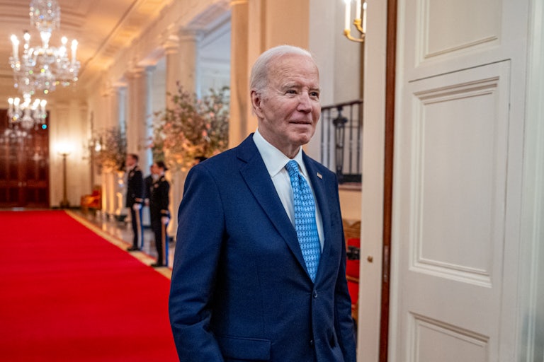 Joe Biden walking. Chandeliers and a red carpet are in the background.