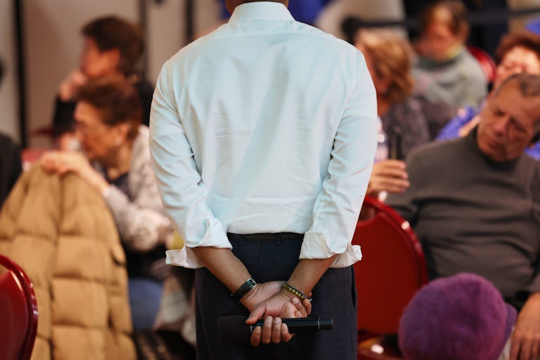 New York City Eric Adams puts both hands behind his back, as he holds a microphone. His head is cut off in the photo.