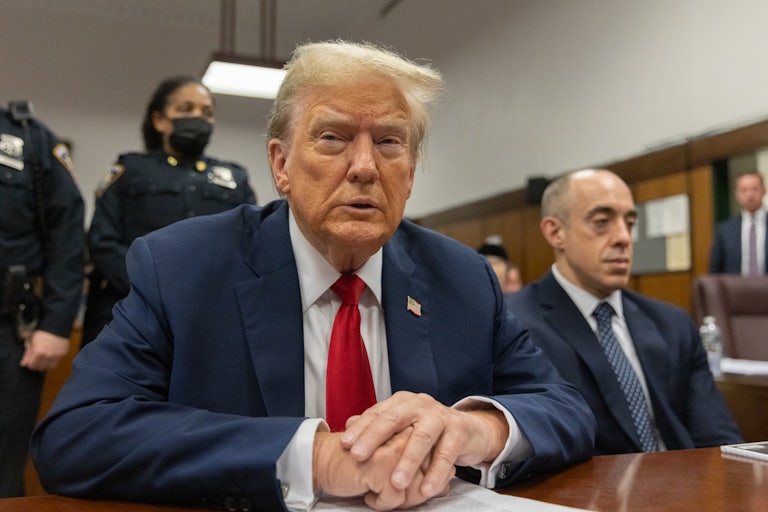 Donald Trump squints as he sits in the courtroom. Two security guards are behind him, and his lawyer is seated on his left.