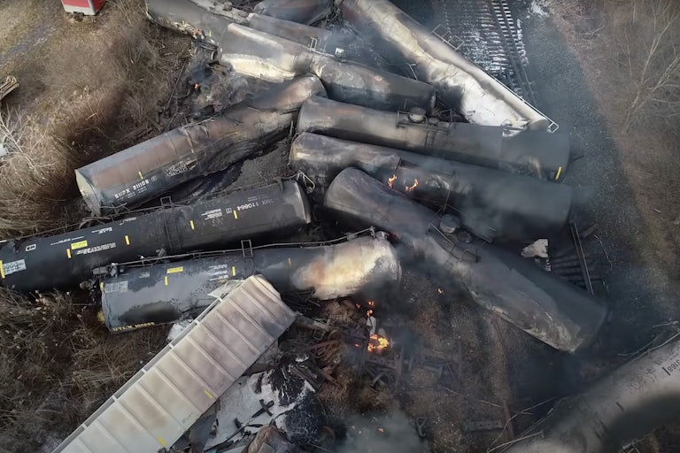 An aerial shot of 11 cargo trains are jammed into one another in a zig-zag pattern. Smoke billows.