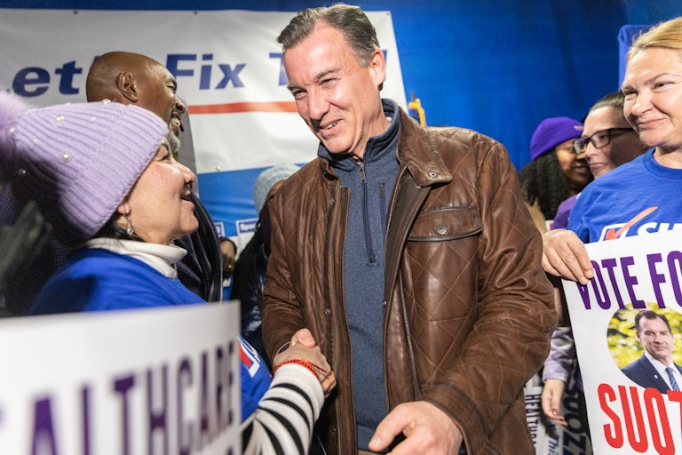 Tom Suozzi smiles and shakes the hand of a supporter. Others surround him and hold his campaign posters.
