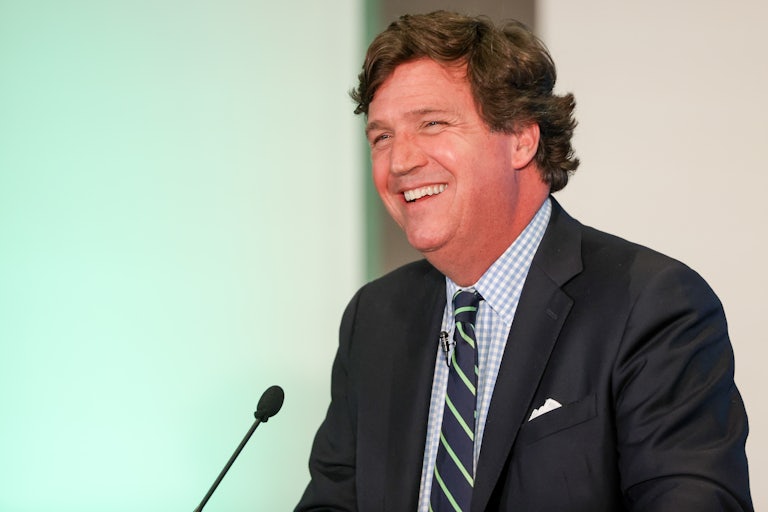 Tucker Carlson, seated, wears a suit and laughs before a mic