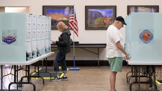 Voters cast their ballots at Desert Vista Community Center in Las Vegas, Nevada. In the foreground to the right, an older white man wearing blue shorts, a white tshirt, and a black cap points at a paper on the table in front of him. In the background to the left, a woman with strawberry blonde hair wearing jeans and a black hoodie looks at a ballot. The booth panel has a sticker that reads "Clark County, Nevada."