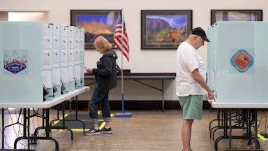 Voters cast their ballots at Desert Vista Community Center in Las Vegas, Nevada. In the foreground to the right, an older white man wearing blue shorts, a white tshirt, and a black cap points at a paper on the table in front of him. In the background to the left, a woman with strawberry blonde hair wearing jeans and a black hoodie looks at a ballot. The booth panel has a sticker that reads "Clark County, Nevada."