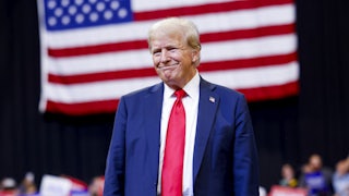 Donald Trump toothlessly smiles as he stands before a large American flag at a political rally
