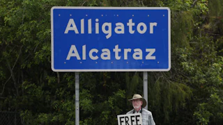 An activist holds a sign that reads "Free Them" as he stands beneath the "Alligator Alcatraz" sign.