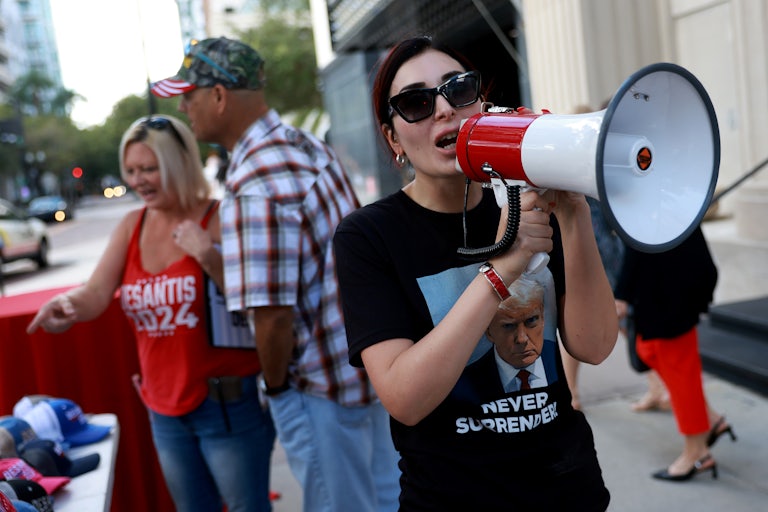 Laura Loomer speaks into a megaphone. She is wearing sunglasses and a shirt with Trump's mugshot and the words "Never Surrender."