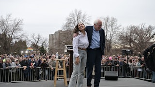 Bernie Sanders puts his arm around Alexandria Ocasio Cortez’s shoulders as both laugh on stage. A huge crowd appears behind them.