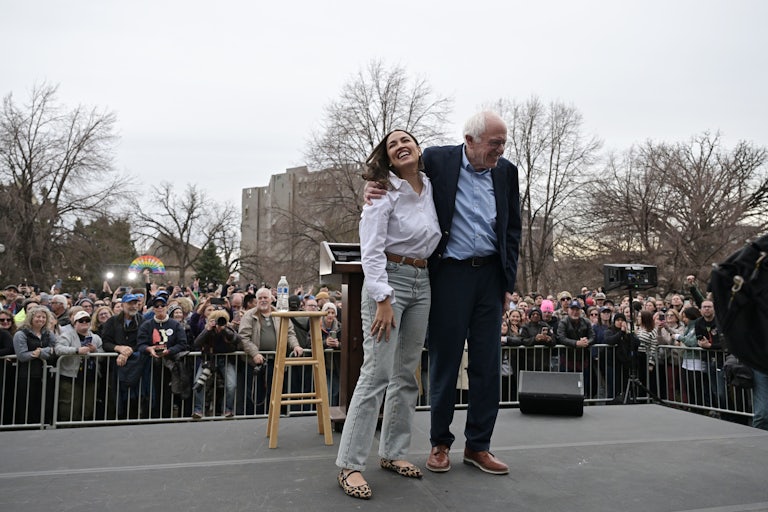 Bernie Sanders puts his arm around Alexandria Ocasio Cortez’s shoulders as both laugh on stage. A huge crowd appears behind them.