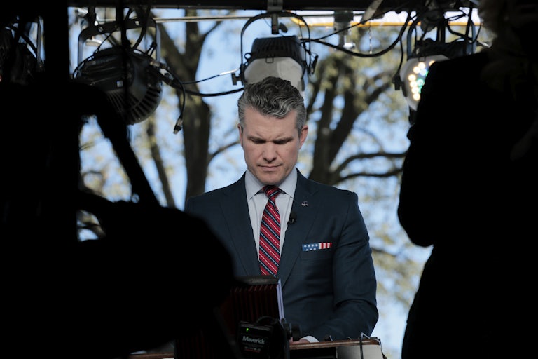 Defense Secretary Pete Hegseth lowers his head looking at a screen as press cameras surround him.