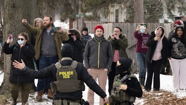 Observers watch and film masked ICE agents in the sidewalk.