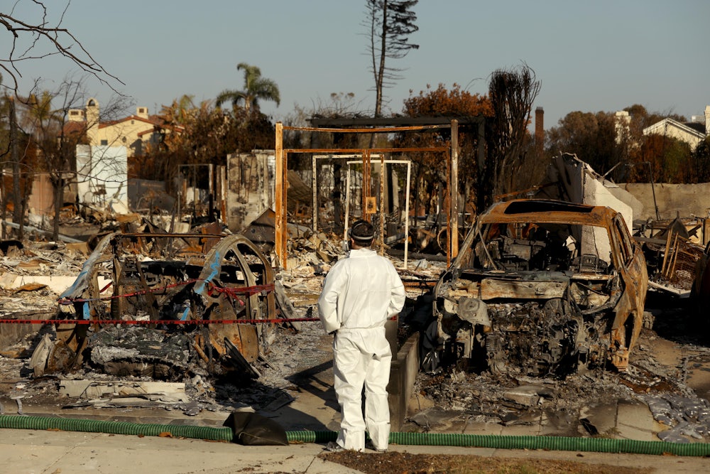 A person in white protective gear stands in front of burned out cars and ash-filled lots.