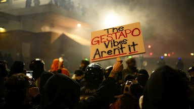 A protester in a crowd holds a sign reading "The Gestapo Vibes Aren't It."