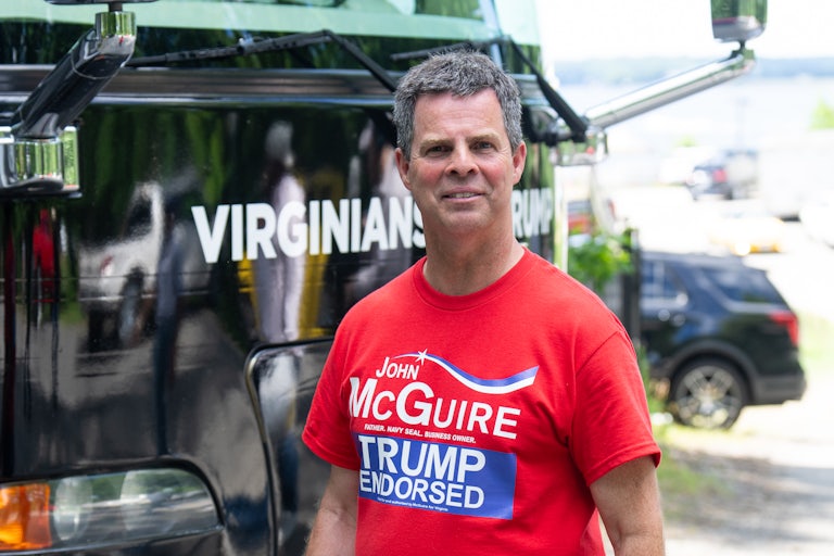 John McGuire stands in front of a vehicle wearing a red shirt reading "McGuire" and "Trump Endorsed."