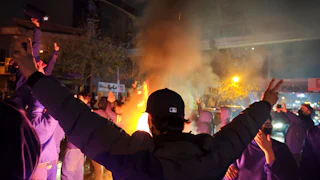 A man in Tehran holds up two peace signs as a fire burns in front of him.