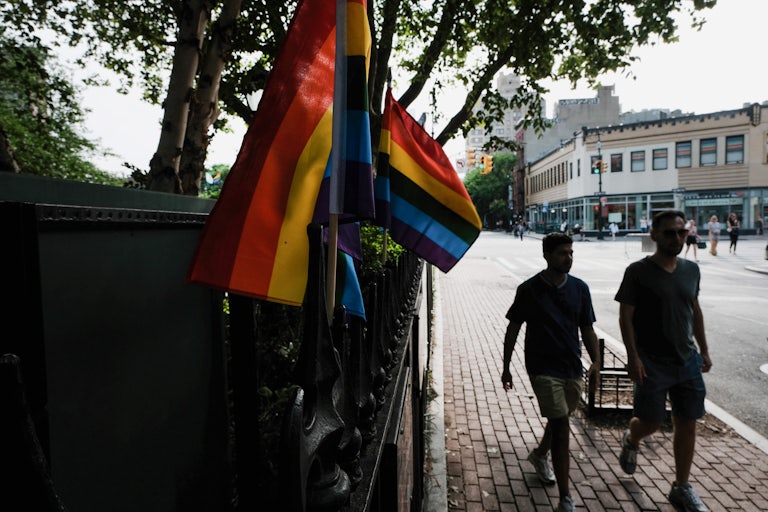 Two people walk by a railing with several pride flags on it