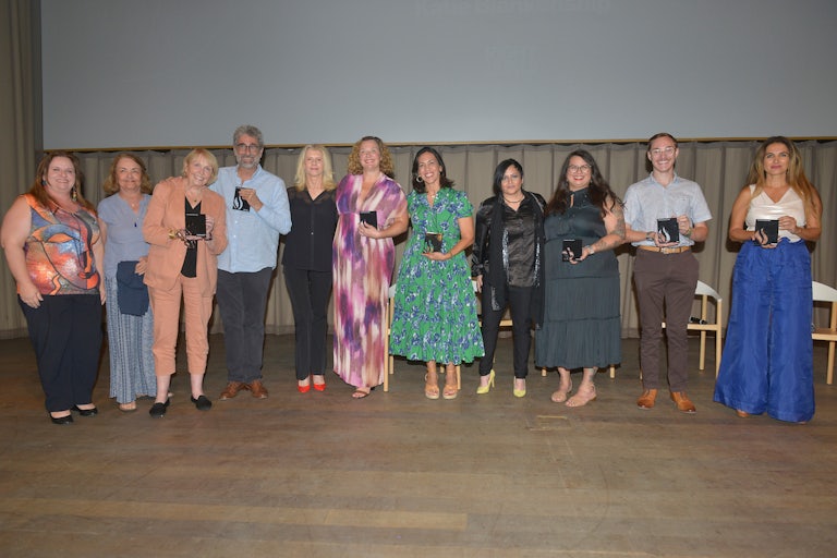 Eleven authors hold up prizes while standing on a stage side by side