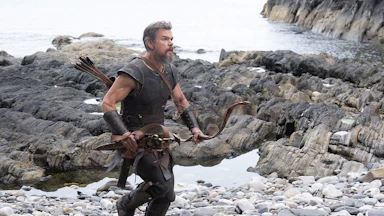 Matt Damon as Odysseus walking over a rocky shoreline in Christopher Nolan’s “The Odyssey”