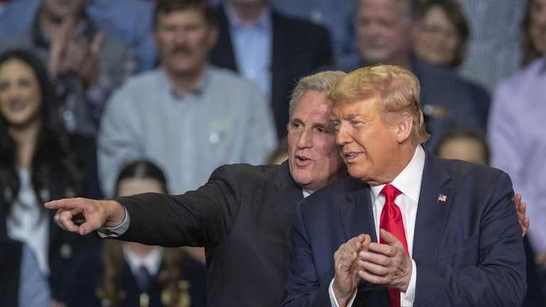 Kevin McCarthy wraps his arm around Donald Trump's shoulders and points off into the distance. They both smile. Trump is clapping. A crowd is in the background
