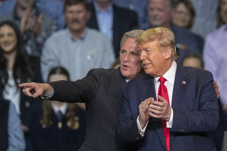 Kevin McCarthy wraps his arm around Donald Trump's shoulders and points off into the distance. They both smile. Trump is clapping. A crowd is in the background