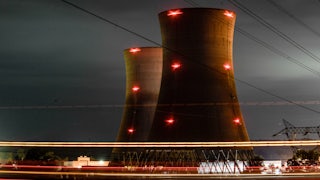Two nuclear cooling towers rise up from the ground at night with red lights on them and power lines in the foreground.