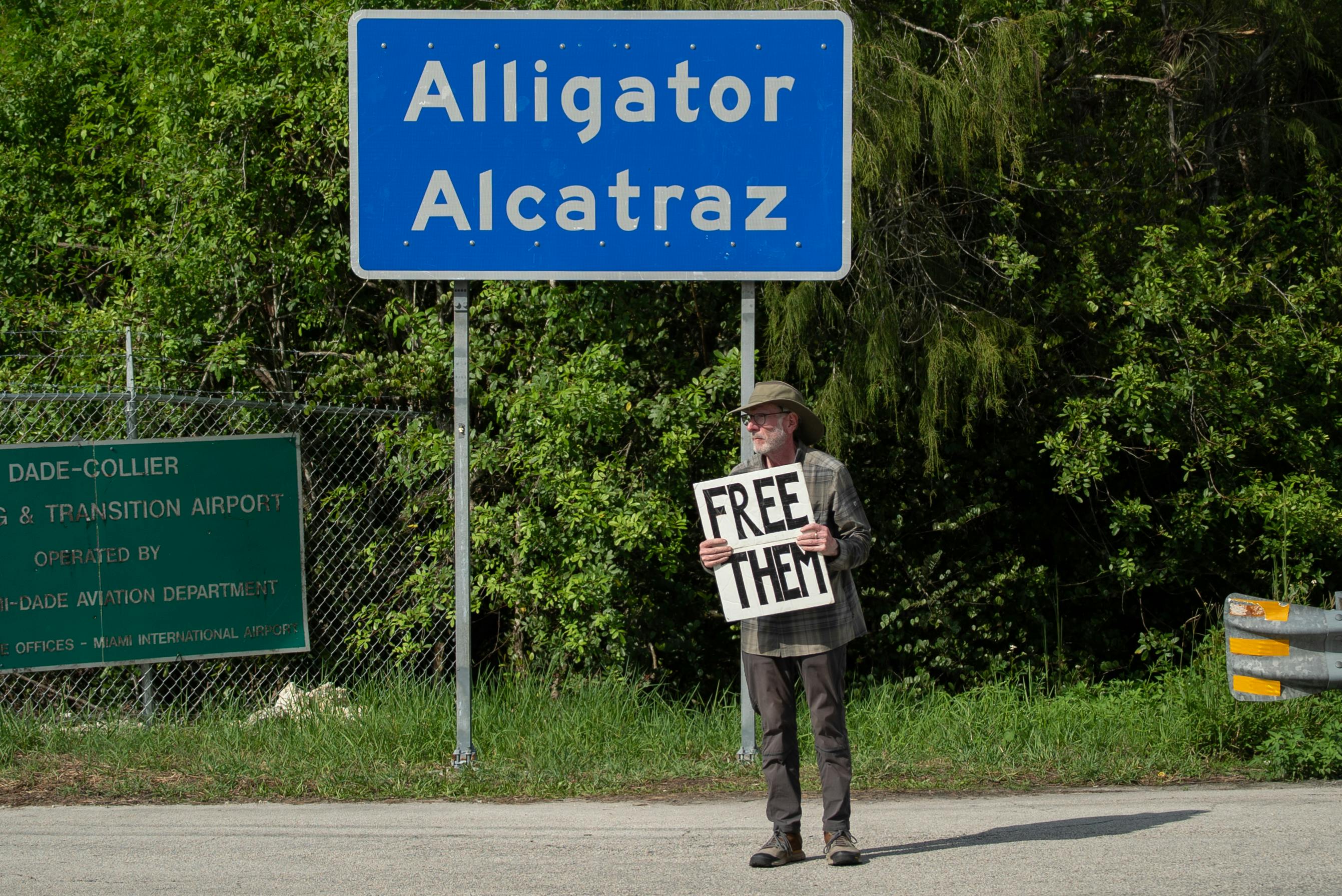 A person holds a sign that says, "Free them" while standing in front of the sign for Alligator Alcatraz