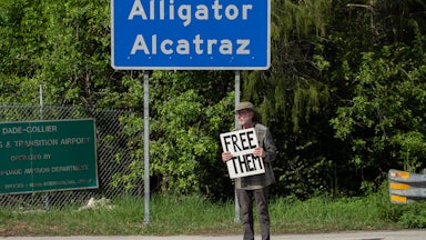 A person holds a sign that says, "Free them" while standing in front of the sign for Alligator Alcatraz