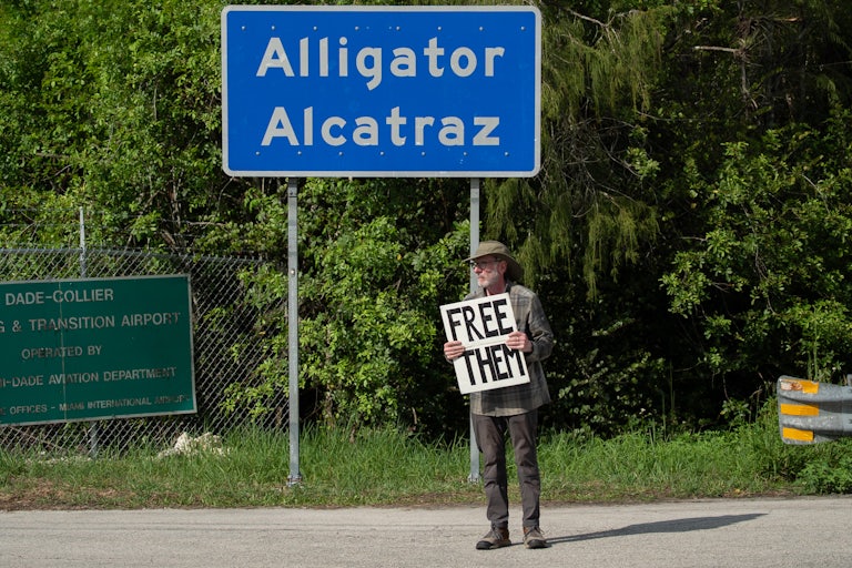 A person holds a sign that says, "Free them" while standing in front of the sign for Alligator Alcatraz
