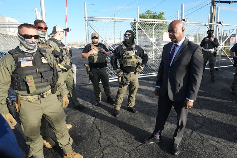 Mayor Ras Baraka of Newark, wearing a suit, confronts several ICE agents wearing protective gear and balaclavas hiding their faces.
