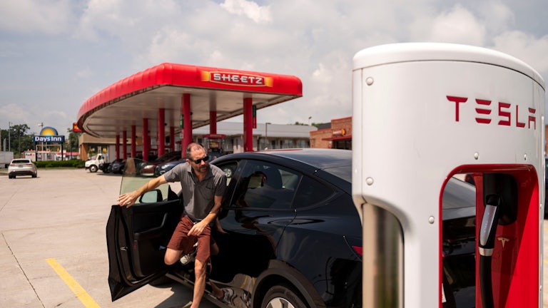 A man exits a Tesla and heads towards a charger in the foreground. In the background, a stand of gas pumps with the Sheetz logo can be seen.