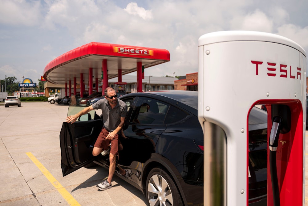 A man exits a Tesla and heads towards a charger in the foreground. In the background, a stand of gas pumps with the Sheetz logo can be seen.