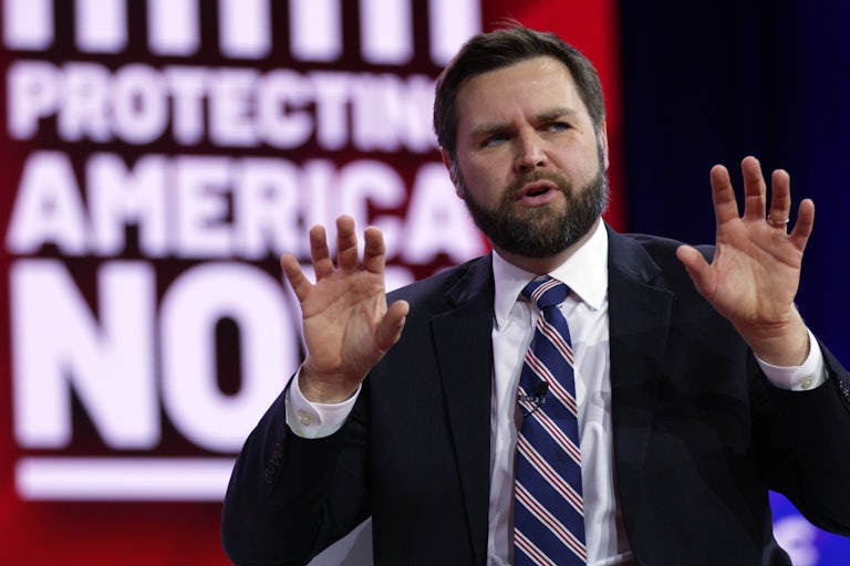 J.D. Vance speaking and holding both hands in the air. The background reads "Protect America Now," out of focus.