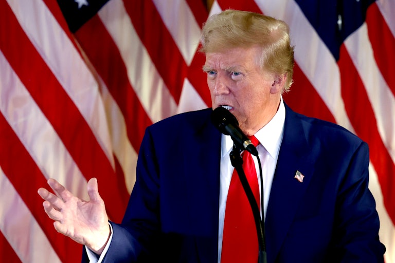 Donald Trump speaks with a mic before him and he gestures with his right hand. There are several U.S flags behind him.