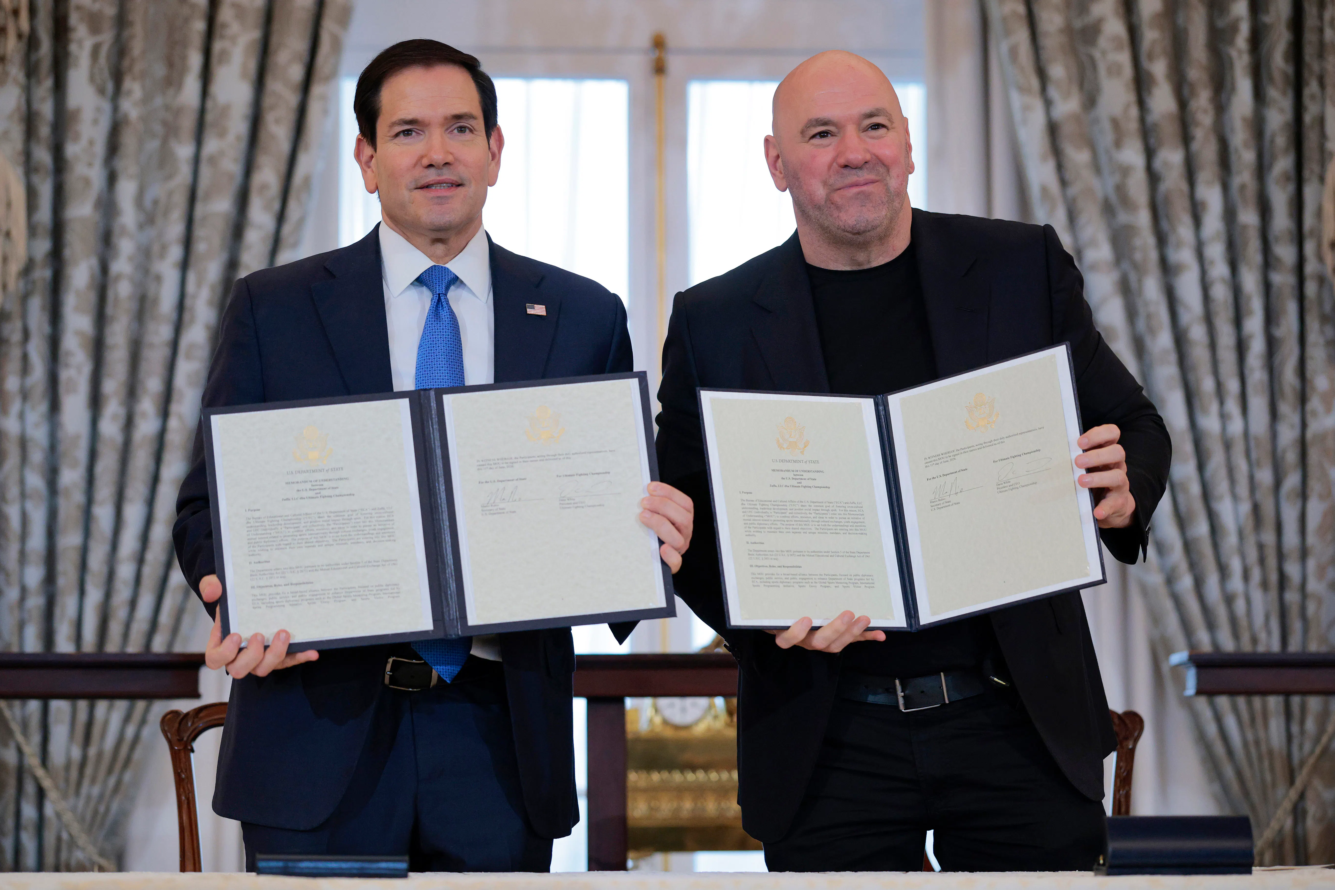 Secretary of State Marco Rubio and Ultimate Fighting Championship CEO Dana White stand next to each other smiling and holding up two copies of a signed memorandum of understanding.