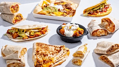 A variety of Taco Bell menu items, from burritos to loaded nachos and other dishes, are seen arrayed on a white background.