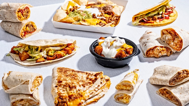 A variety of Taco Bell menu items, from burritos to loaded nachos and other dishes, are seen arrayed on a white background.