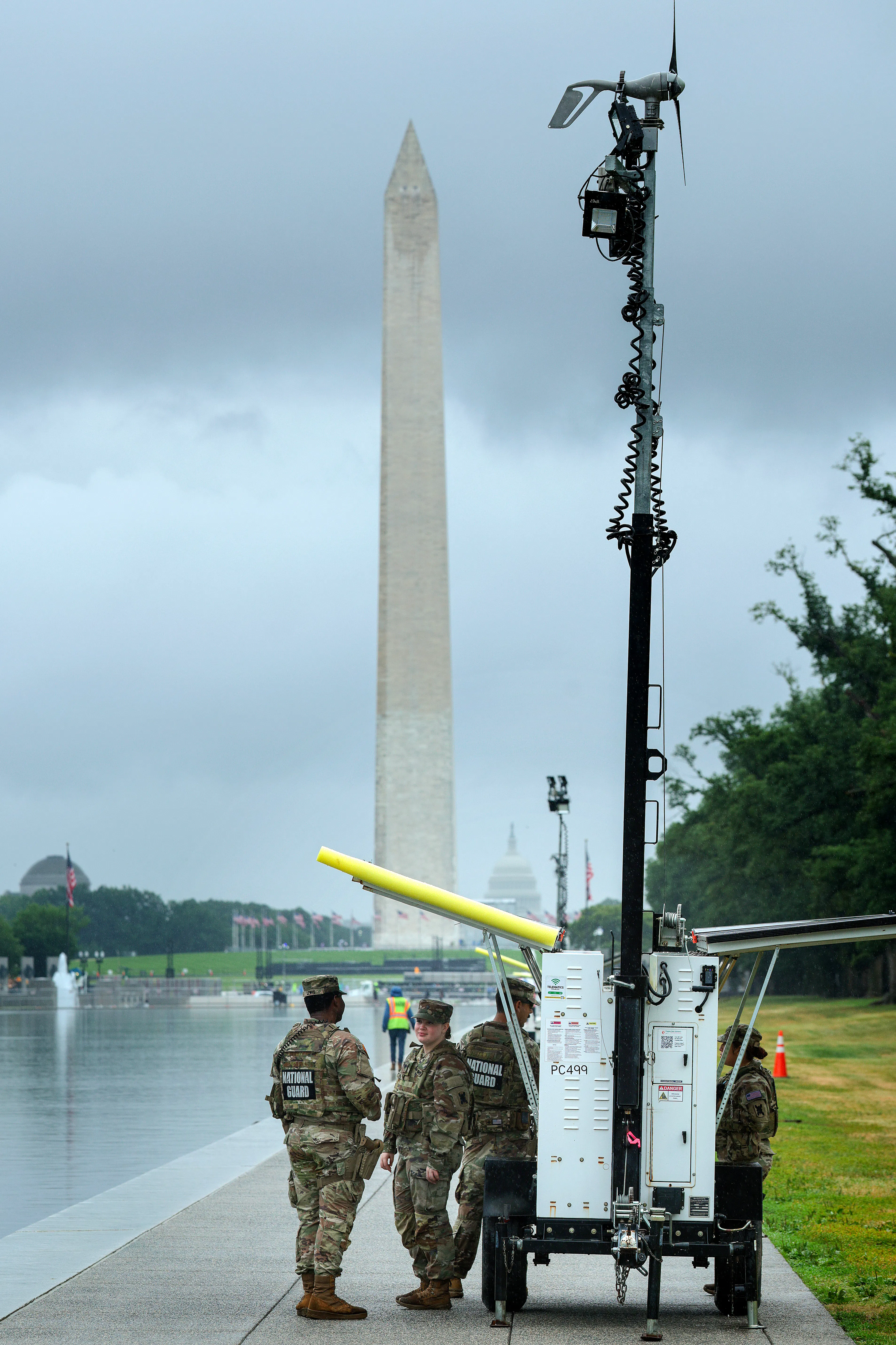 U.S. Army National Guard troops take shelter from the rain underneath a mobile camera trailer’s solar panels while guarding the Lincoln Memorial Reflecting Pool. The Washington Monument is in the background.