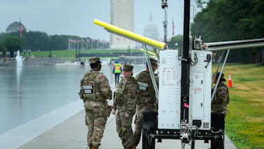 U.S. Army National Guard troops take shelter from the rain underneath a mobile camera trailer’s solar panels while guarding the Lincoln Memorial Reflecting Pool. The Washington Monument is in the background.