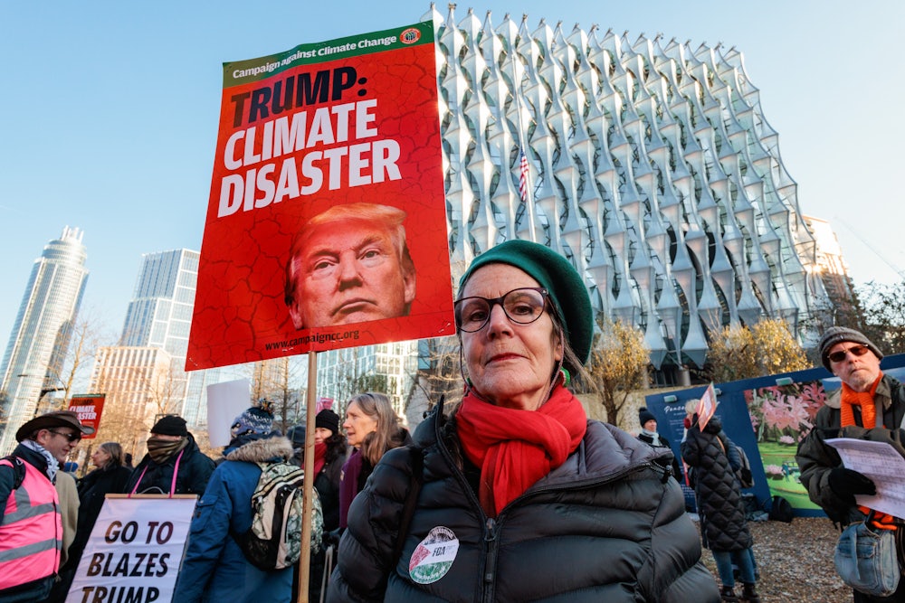 A climate activist wearing a red scarf holds a red placard reading "Trump: Climate Disaster," with other activists in the background.