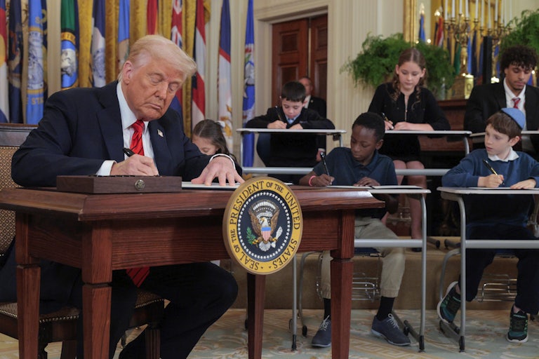 Donald Trump signs an executive order to shrink the Education Department alongside school children signing their own versions in a White House ceremony. They all sit at their own desks, Trump included.