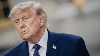Donald Trump, wearing a blue suit and blue tie, stands at an angle.