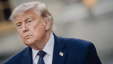 Donald Trump, wearing a blue suit and blue tie, stands at an angle.