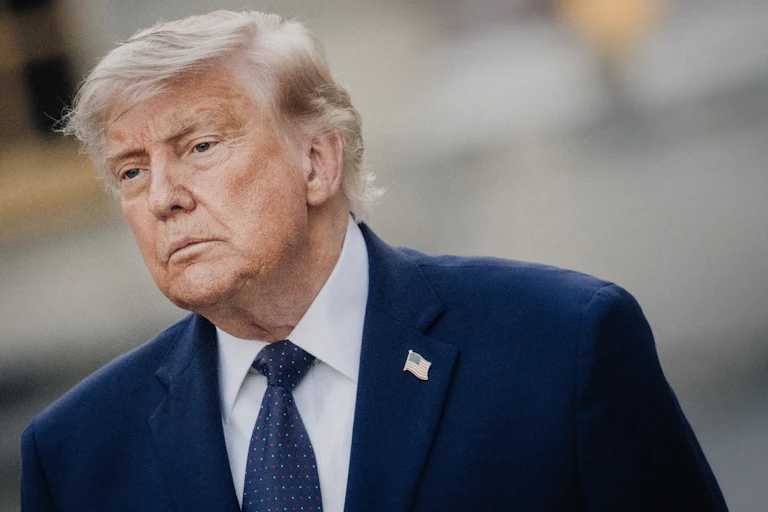 Donald Trump, wearing a blue suit and blue tie, stands at an angle.